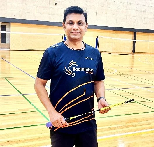 Coach Kiran holding a badminton racket on the indoor court, from his coach profile